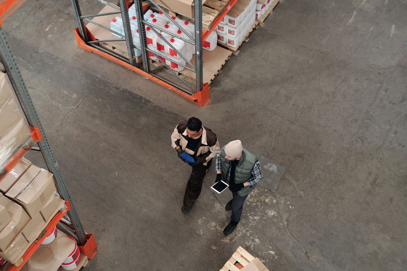 Vista de cima de duas pessoas a conversar, uma com um tablet na mão, no corredor de um armazém industrial